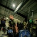 A person in a military-style olive green t-shirt is performing a deadlift with a heavily loaded barbell in an indoor gym facility. The barbell is loaded with Hammer Strength 45-lb blue bumper plates on both sides. Several onlookers, some in military camouflage uniforms, observe the lift in the background.