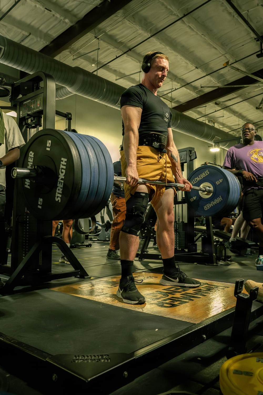 A muscular man is performing a deadlift on a raised platform in a commercial gym. He is wearing a weightlifting belt, knee brace, black t-shirt, mustard-yellow shorts, and over-ear headphones. The barbell is loaded with multiple Hammer Strength plates on each side.