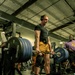 A muscular man is performing a deadlift on a raised platform in a commercial gym. He is wearing a weightlifting belt, knee brace, black t-shirt, mustard-yellow shorts, and over-ear headphones. The barbell is loaded with multiple Hammer Strength plates on each side.