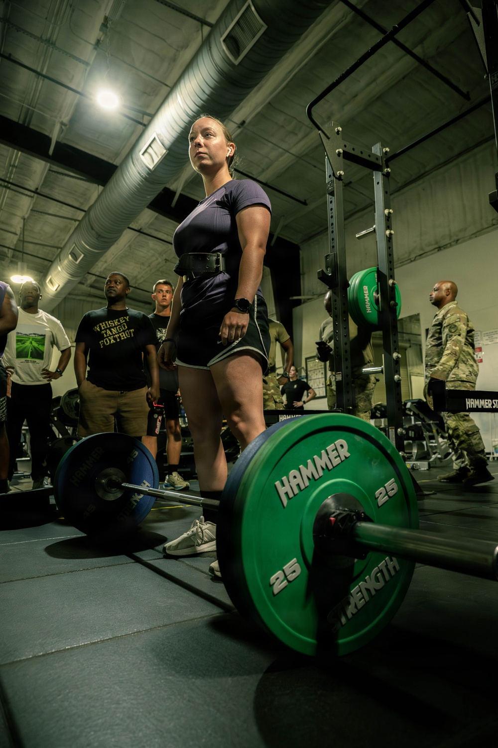 A woman wearing a weightlifting belt, dark athletic clothing, and a smartwatch stands at a loaded barbell in an indoor gym facility. She appears to be preparing for or has just completed a deadlift, with spectators including individuals in military camouflage uniforms watching in the background. The barbell is loaded with Hammer Strength 25 lb green bumper plates.