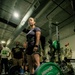 A woman wearing a weightlifting belt, dark athletic clothing, and a smartwatch stands at a loaded barbell in an indoor gym facility. She appears to be preparing for or has just completed a deadlift, with spectators including individuals in military camouflage uniforms watching in the background. The barbell is loaded with Hammer Strength 25 lb green bumper plates.