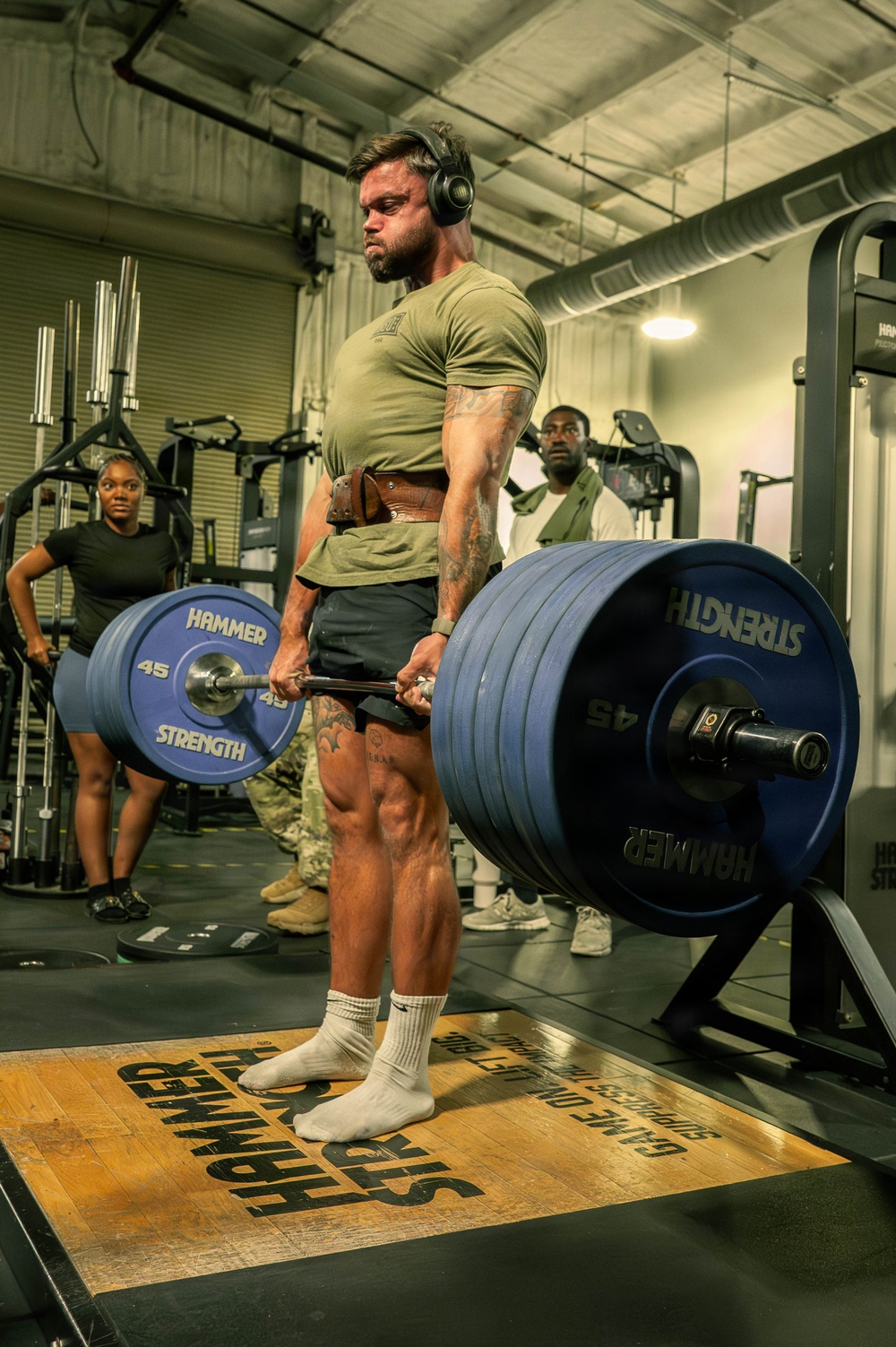 A heavily muscular man performs a deadlift in a gym equipped with Hammer Strength equipment. He is wearing a olive green t-shirt, black shorts, white socks, a leather weightlifting belt, and over-ear headphones. Two other individuals are visible in the background observing or using other equipment.