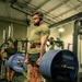 A heavily muscular man performs a deadlift in a gym equipped with Hammer Strength equipment. He is wearing a olive green t-shirt, black shorts, white socks, a leather weightlifting belt, and over-ear headphones. Two other individuals are visible in the background observing or using other equipment.