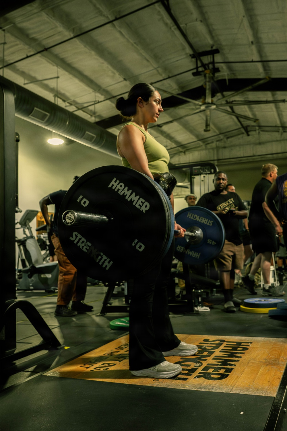 A woman in a green tank top and black pants performs a deadlift in a gym setting, holding a barbell loaded with Hammer Strength bumper plates. The environment appears to be a well-equipped commercial or specialty gym with multiple people visible in the background. The floor mat beneath her is branded with 'Hammer Strength' text.