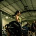 A woman in a green tank top and black pants performs a deadlift in a gym setting, holding a barbell loaded with Hammer Strength bumper plates. The environment appears to be a well-equipped commercial or specialty gym with multiple people visible in the background. The floor mat beneath her is branded with 'Hammer Strength' text.