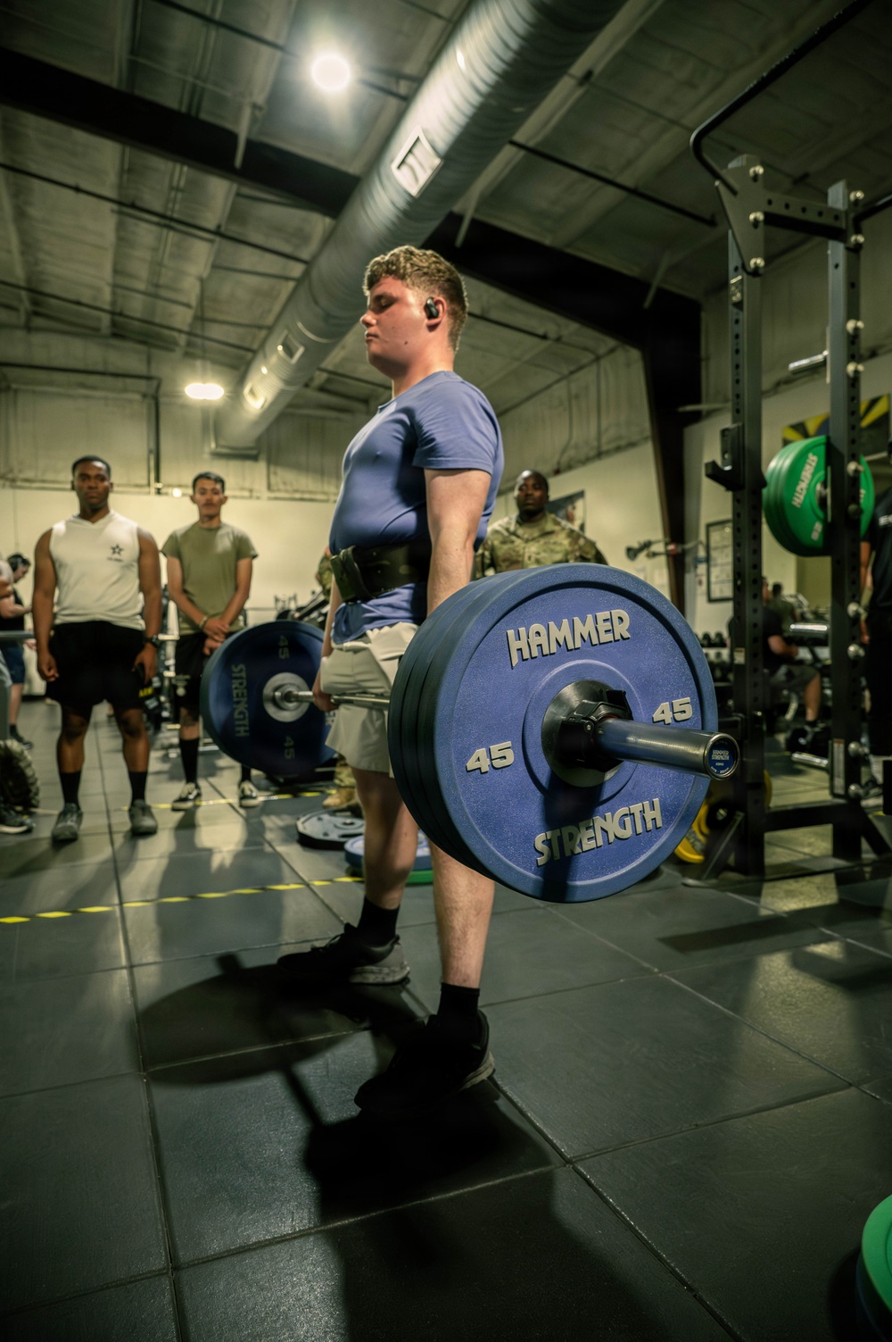 A young male athlete is performing a deadlift in an indoor gym facility, holding a loaded barbell with Hammer Strength 45-lb bumper plates. He is wearing a blue t-shirt, shorts, black socks, and a weightlifting belt, with wireless earbuds in. Several observers, including individuals in military-style clothing, are watching in the background.