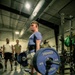 A young male athlete is performing a deadlift in an indoor gym facility, holding a loaded barbell with Hammer Strength 45-lb bumper plates. He is wearing a blue t-shirt, shorts, black socks, and a weightlifting belt, with wireless earbuds in. Several observers, including individuals in military-style clothing, are watching in the background.