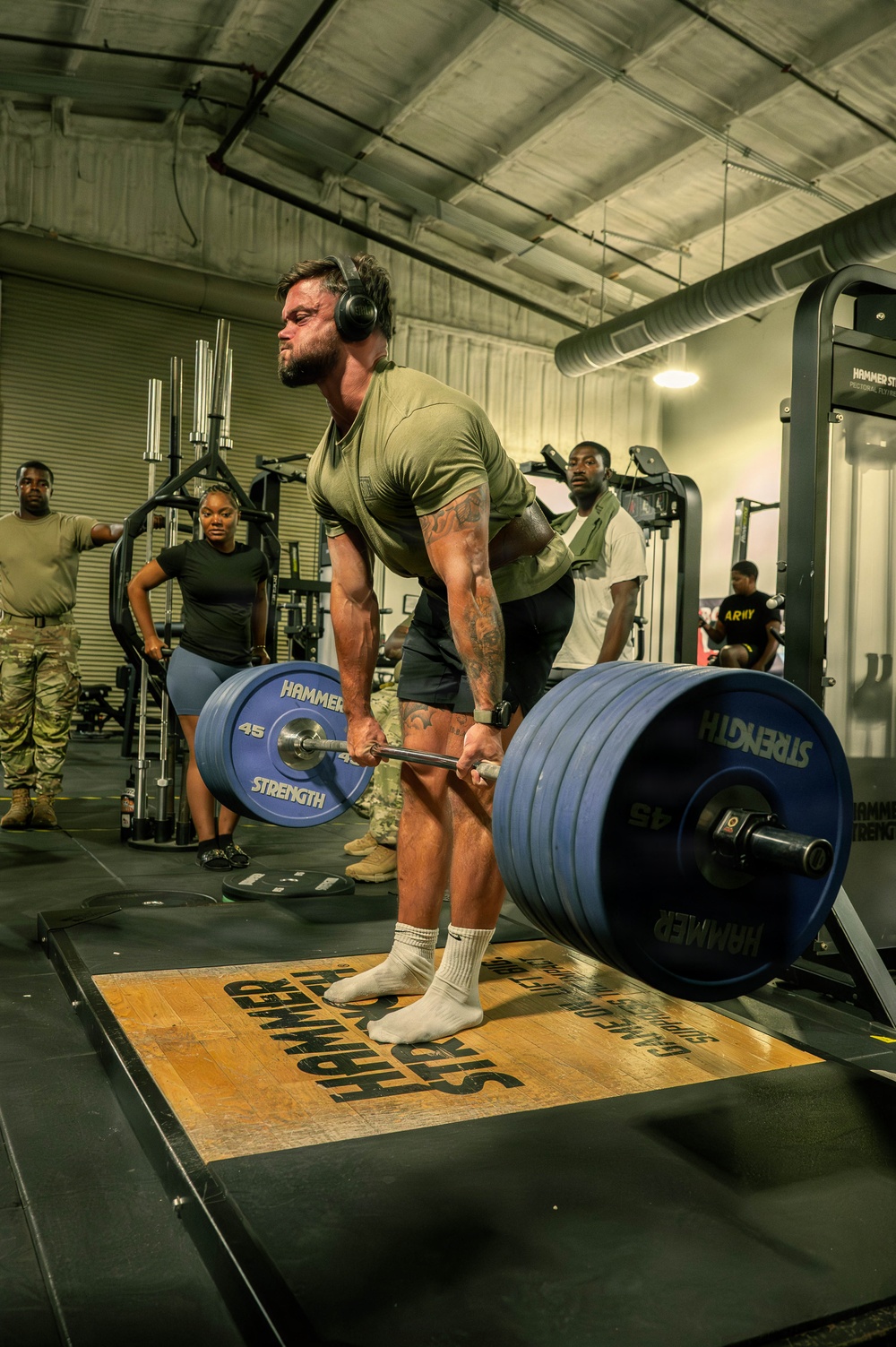 A muscular man wearing a green t-shirt, black shorts, and headphones performs a deadlift on a Hammer Strength platform inside a military gym facility. Multiple observers, some in military camouflage uniforms, watch from the background. The barbell is loaded with multiple 45 lb Hammer Strength plates on each side.