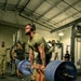 A muscular man wearing a green t-shirt, black shorts, and headphones performs a deadlift on a Hammer Strength platform inside a military gym facility. Multiple observers, some in military camouflage uniforms, watch from the background. The barbell is loaded with multiple 45 lb Hammer Strength plates on each side.