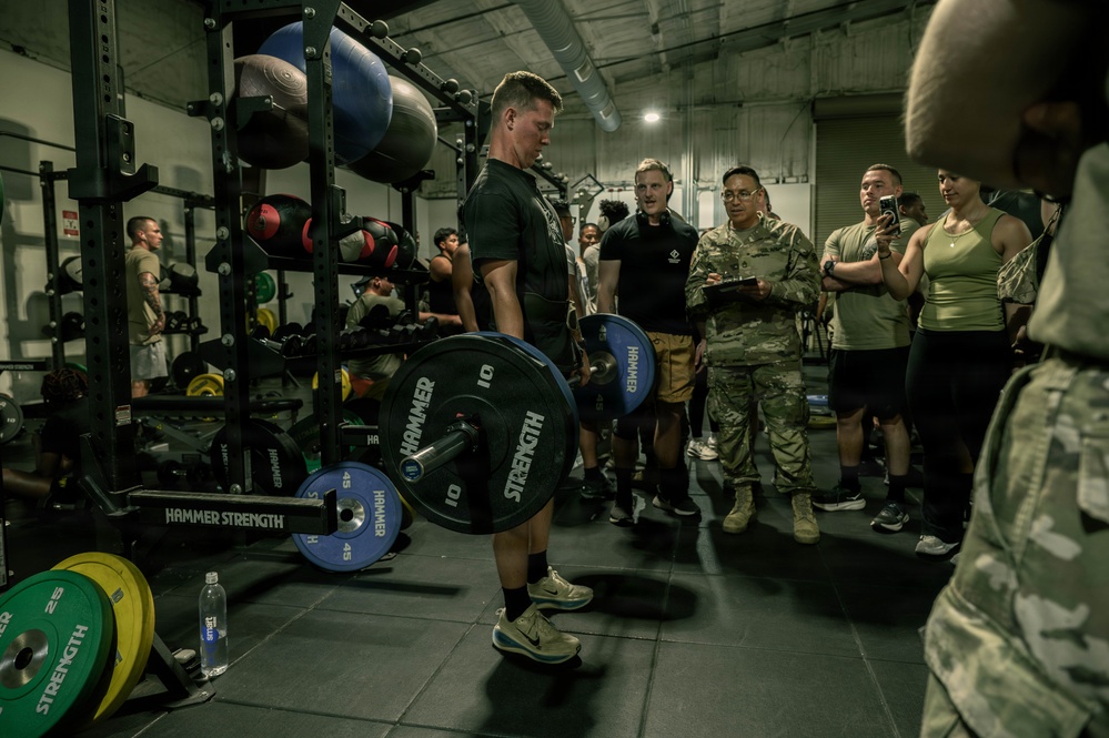A military fitness event is taking place inside a gym equipped with Hammer Strength equipment. A male athlete in a black t-shirt is performing a deadlift while a crowd of military personnel and civilians observe, with at least one person recording on a smartphone. The setting appears to be a structured competition or fitness assessment.