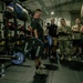A military fitness event is taking place inside a gym equipped with Hammer Strength equipment. A male athlete in a black t-shirt is performing a deadlift while a crowd of military personnel and civilians observe, with at least one person recording on a smartphone. The setting appears to be a structured competition or fitness assessment.