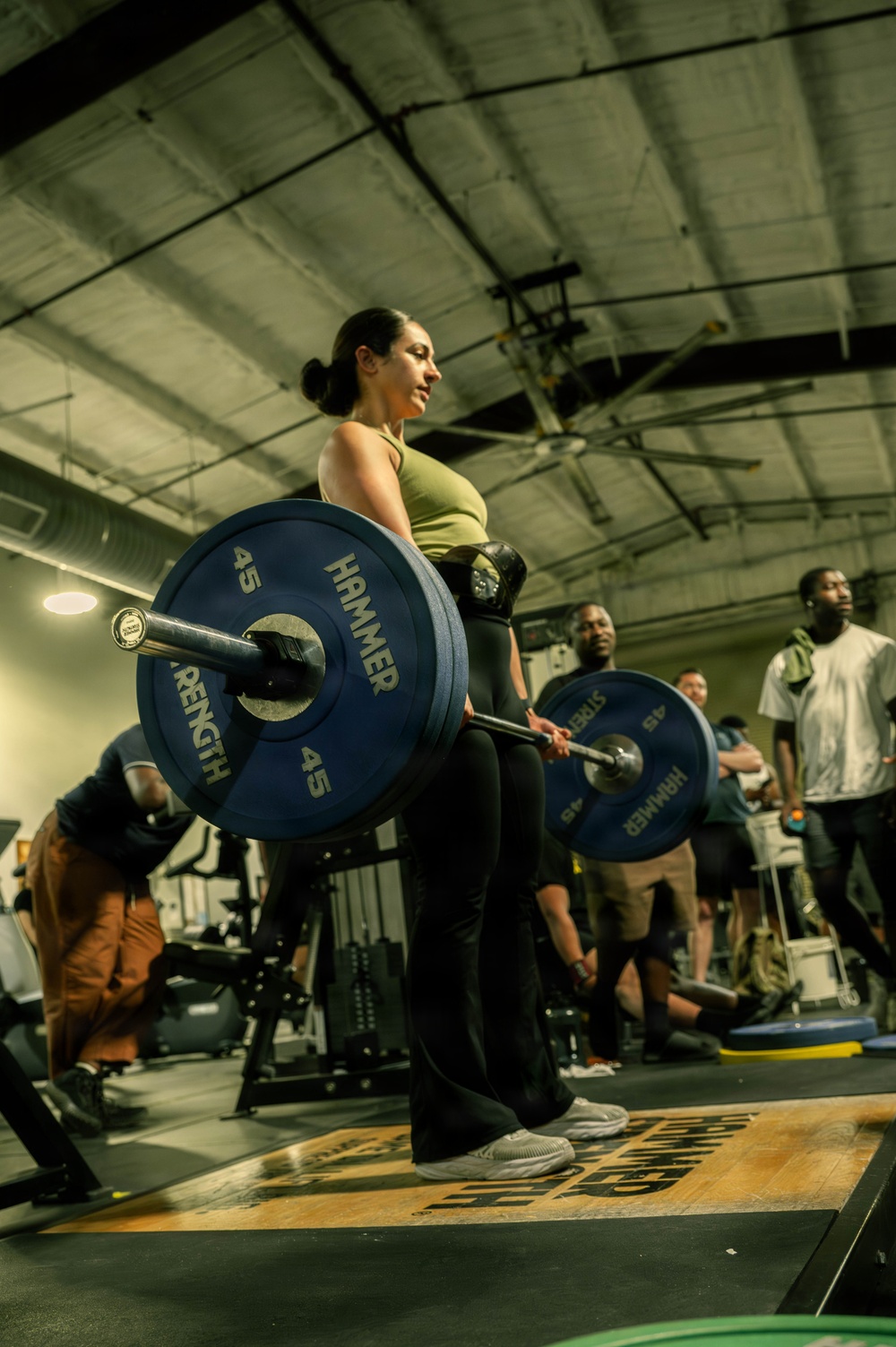 A woman in a green sports top and black leggings performs a deadlift with a heavily loaded barbell in a gym setting. The barbell is loaded with Hammer Strength 45-lb bumper plates on each side. Several onlookers and other gym-goers are visible in the background, suggesting a group training session or competition event.