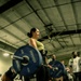 A woman in a green sports top and black leggings performs a deadlift with a heavily loaded barbell in a gym setting. The barbell is loaded with Hammer Strength 45-lb bumper plates on each side. Several onlookers and other gym-goers are visible in the background, suggesting a group training session or competition event.