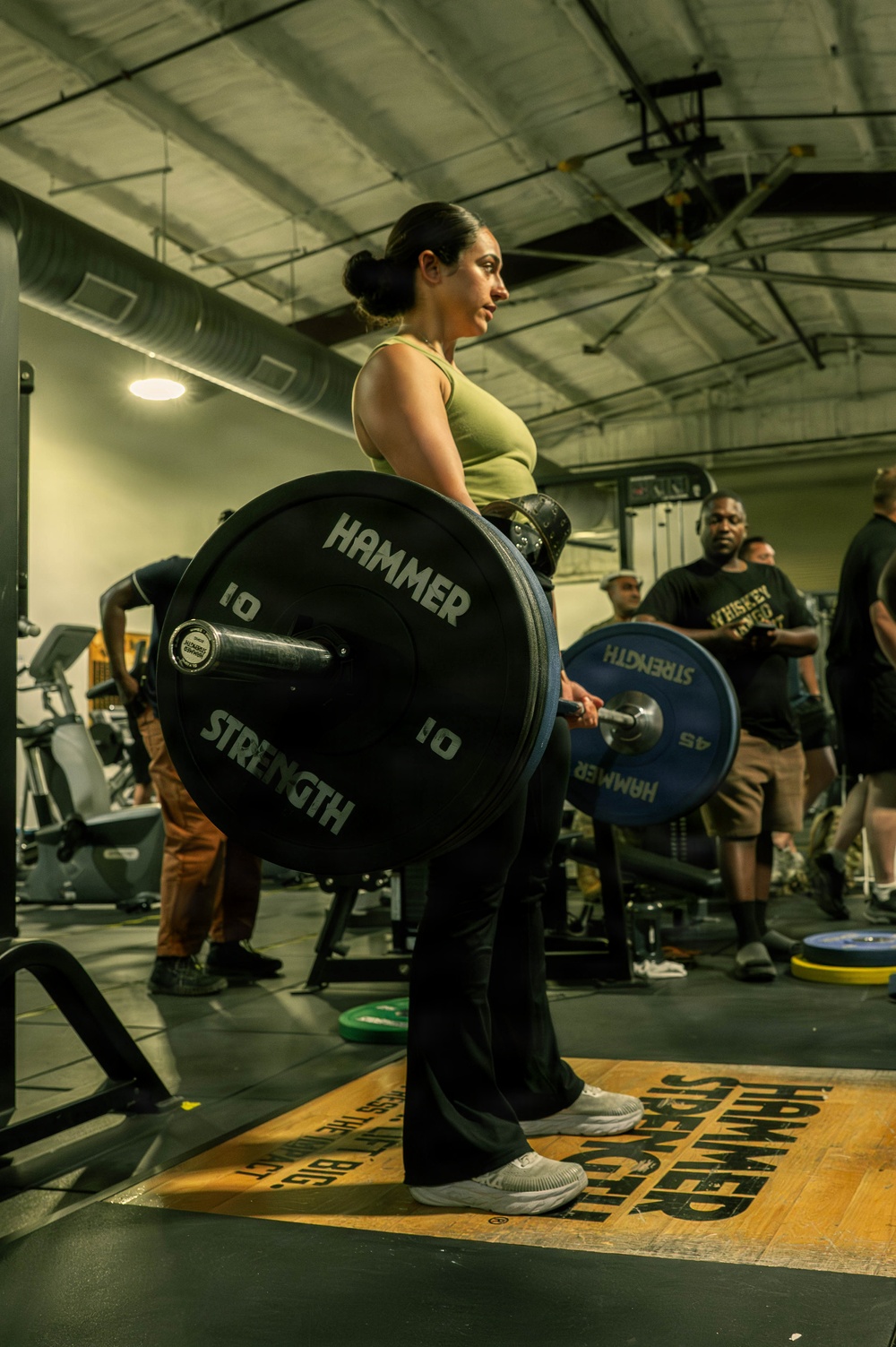 A woman is performing a deadlift in a gym, holding a loaded barbell with Hammer Strength bumper plates. She is standing on a Hammer Strength branded lifting platform, wearing a green tank top, black pants, and white sneakers with a weightlifting belt. Several onlookers are visible in the background, suggesting a gym event or competition setting.