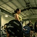 A woman is performing a deadlift in a gym, holding a loaded barbell with Hammer Strength bumper plates. She is standing on a Hammer Strength branded lifting platform, wearing a green tank top, black pants, and white sneakers with a weightlifting belt. Several onlookers are visible in the background, suggesting a gym event or competition setting.