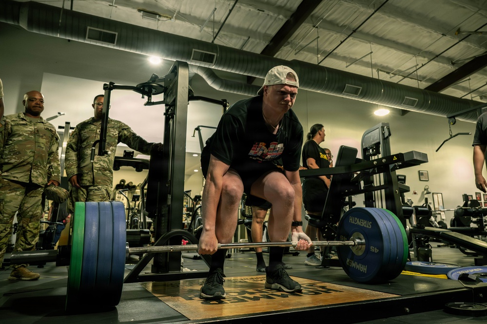 A person in civilian athletic wear is performing a deadlift with a heavily loaded barbell in a military gym facility. Several individuals in U.S. Army camouflage (OCP) uniforms are observing from the background. The setting appears to be an official military fitness center, possibly during a competition or demonstration event.