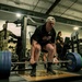 A person in civilian athletic wear is performing a deadlift with a heavily loaded barbell in a military gym facility. Several individuals in U.S. Army camouflage (OCP) uniforms are observing from the background. The setting appears to be an official military fitness center, possibly during a competition or demonstration event.