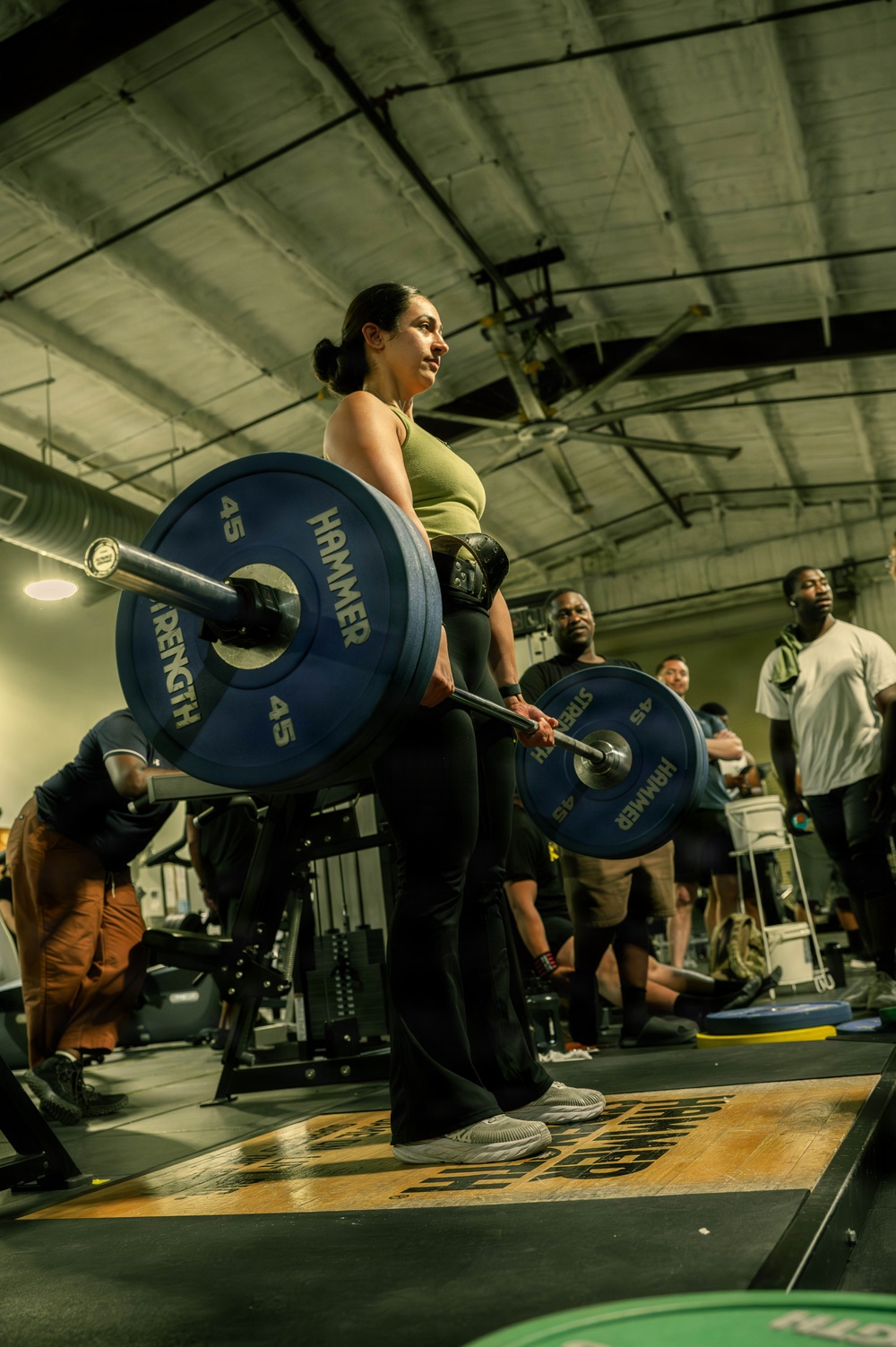 A woman is performing a deadlift in a gym setting, holding a loaded barbell with Hammer Strength 45-lb bumper plates on each side. She is wearing a green tank top, black pants, a lifting belt, and gray sneakers, and appears to be mid-lift or at lockout position. Several onlookers and other gym-goers are visible in the background.