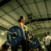 A woman is performing a deadlift in a gym setting, holding a loaded barbell with Hammer Strength 45-lb bumper plates on each side. She is wearing a green tank top, black pants, a lifting belt, and gray sneakers, and appears to be mid-lift or at lockout position. Several onlookers and other gym-goers are visible in the background.