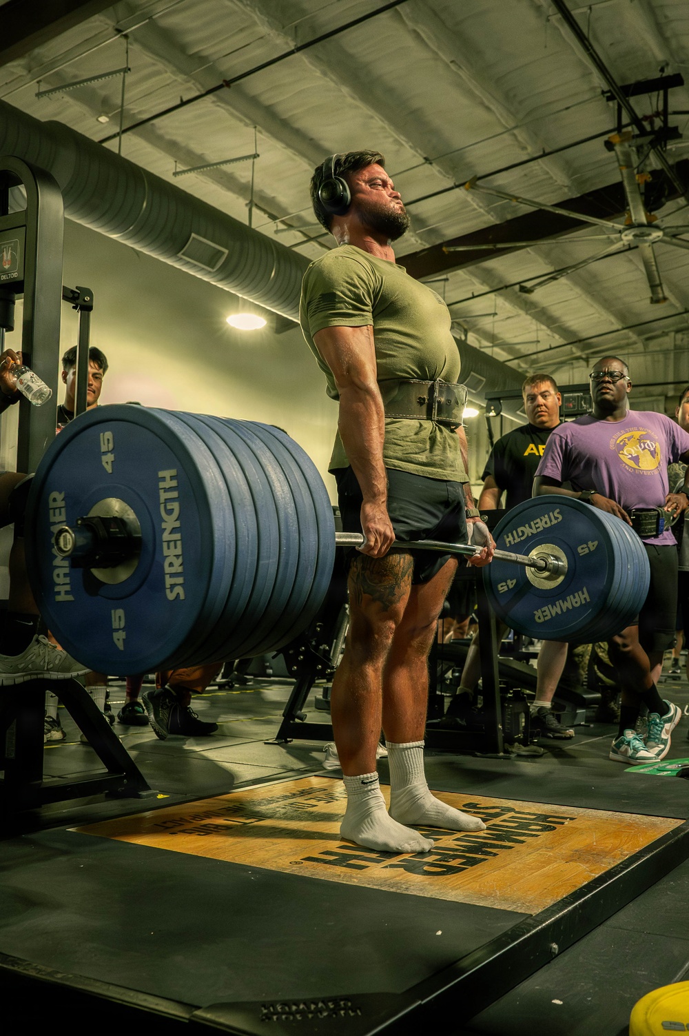 A muscular male athlete is performing a heavy deadlift in a gym setting, wearing a weightlifting belt, headphones, and a green t-shirt. The barbell is loaded with multiple Hammer Strength 45-lb plates on each side. Several spectators and other gym-goers are visible in the background, suggesting a competitive or group training event.
