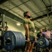 A muscular male athlete is performing a heavy deadlift in a gym setting, wearing a weightlifting belt, headphones, and a green t-shirt. The barbell is loaded with multiple Hammer Strength 45-lb plates on each side. Several spectators and other gym-goers are visible in the background, suggesting a competitive or group training event.