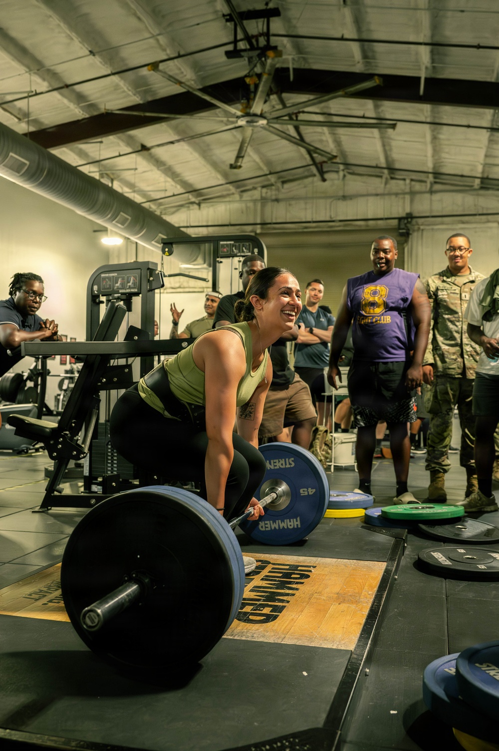 A woman in a green tank top and black leggings is performing a deadlift on a Hammer Strength platform in a gym setting. She is smiling broadly while gripping a loaded barbell with 45 lb plates. A group of onlookers, including individuals in military camouflage uniforms and casual athletic wear, are watching and cheering her on.