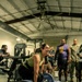 A woman in a green tank top and black leggings is performing a deadlift on a Hammer Strength platform in a gym setting. She is smiling broadly while gripping a loaded barbell with 45 lb plates. A group of onlookers, including individuals in military camouflage uniforms and casual athletic wear, are watching and cheering her on.