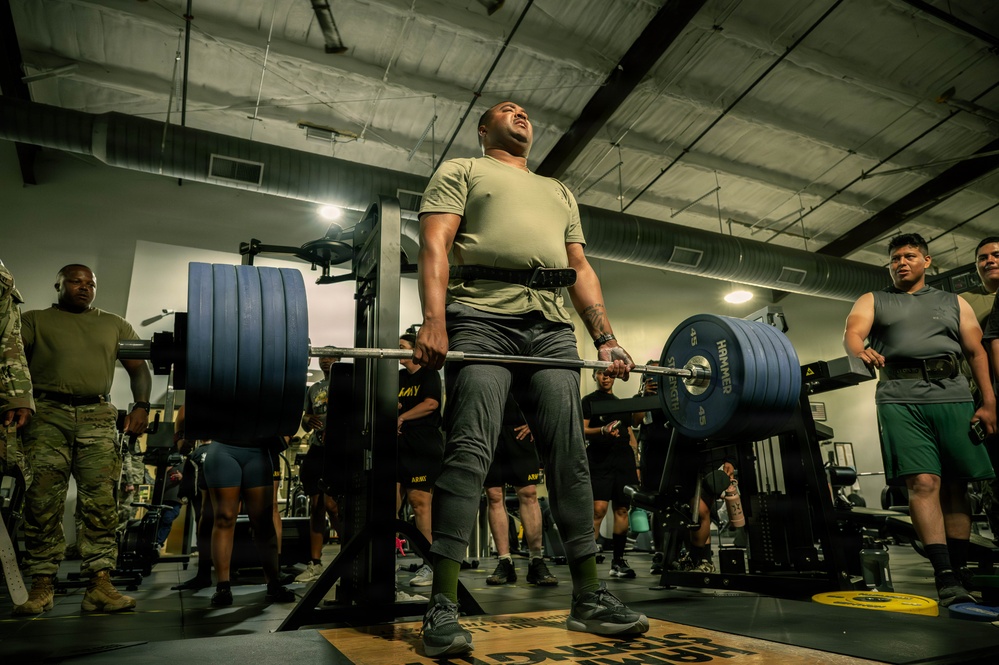 A military service member is performing a heavy deadlift in a gym facility, wearing an olive green Army physical training shirt and a weightlifting belt. Multiple other service members in Army PT gear are visible in the background, suggesting a group physical training session or competition event. The lifter appears to be exerting maximum effort, with head tilted back and an expression of intense strain.