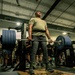 A military service member is performing a heavy deadlift in a gym facility, wearing an olive green Army physical training shirt and a weightlifting belt. Multiple other service members in Army PT gear are visible in the background, suggesting a group physical training session or competition event. The lifter appears to be exerting maximum effort, with head tilted back and an expression of intense strain.