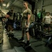 A military fitness assessment is taking place in a gym facility, where a soldier in an olive/tan t-shirt and shorts is performing a deadlift with a Hammer Strength barbell. A uniformed soldier in MultiCam/OCP camouflage with an 82nd Airborne patch and US flag is observing and recording on a clipboard. Several other personnel are visible in the background, suggesting a structured physical fitness test or competition.