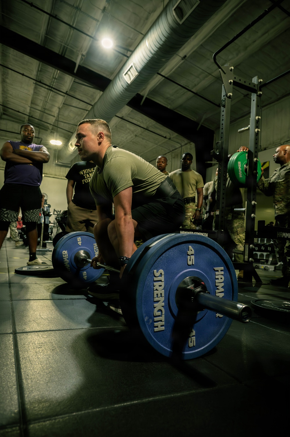 A male athlete in an olive/tan military-style shirt is in the setup position for a deadlift, gripping a loaded barbell with Hammer Strength 45-lb bumper plates. The scene takes place in a military or institutional gym facility with several onlookers, some in military uniforms, observing in the background. The low-angle, dramatic lighting emphasizes the weight plates and the lifter's muscular build.