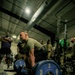 A male athlete in an olive/tan military-style shirt is in the setup position for a deadlift, gripping a loaded barbell with Hammer Strength 45-lb bumper plates. The scene takes place in a military or institutional gym facility with several onlookers, some in military uniforms, observing in the background. The low-angle, dramatic lighting emphasizes the weight plates and the lifter's muscular build.