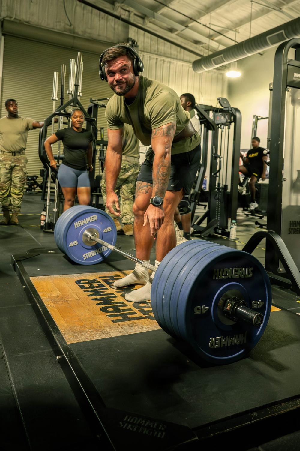 A tattooed male athlete wearing headphones and an olive green t-shirt is positioned over a heavily loaded barbell on a Hammer Strength lifting platform, appearing to be mid-deadlift or preparing for one. Several other individuals in military-style attire observe in the background of what appears to be a military fitness facility. The gym is well-equipped with multiple Hammer Strength machines and racks visible.