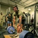 A tattooed male athlete wearing headphones and an olive green t-shirt is positioned over a heavily loaded barbell on a Hammer Strength lifting platform, appearing to be mid-deadlift or preparing for one. Several other individuals in military-style attire observe in the background of what appears to be a military fitness facility. The gym is well-equipped with multiple Hammer Strength machines and racks visible.