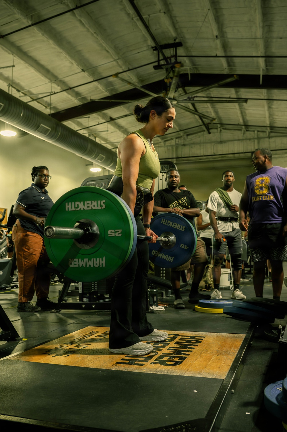 A woman is performing a deadlift in what appears to be a gym competition or training event, standing on a branded Hammer Strength platform. She is lifting a loaded barbell with green and blue weight plates marked 'Hammer Strength.' Several spectators and participants are visible in the background observing the lift.