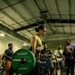 A woman is performing a deadlift in what appears to be a gym competition or training event, standing on a branded Hammer Strength platform. She is lifting a loaded barbell with green and blue weight plates marked 'Hammer Strength.' Several spectators and participants are visible in the background observing the lift.