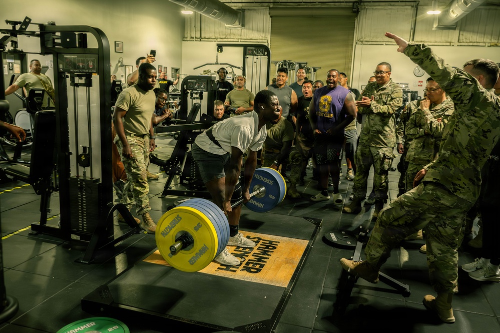 A military gym setting where a soldier in civilian workout clothes is performing a heavy deadlift on a Hammer Strength platform, surrounded by a large crowd of military personnel in camouflage uniforms cheering and reacting enthusiastically. The lifter appears to be straining under significant weight loaded with yellow and blue bumper plates. The atmosphere is energetic and competitive, suggesting an informal powerlifting event or fitness challenge.