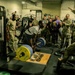 A military gym setting where a soldier in civilian workout clothes is performing a heavy deadlift on a Hammer Strength platform, surrounded by a large crowd of military personnel in camouflage uniforms cheering and reacting enthusiastically. The lifter appears to be straining under significant weight loaded with yellow and blue bumper plates. The atmosphere is energetic and competitive, suggesting an informal powerlifting event or fitness challenge.