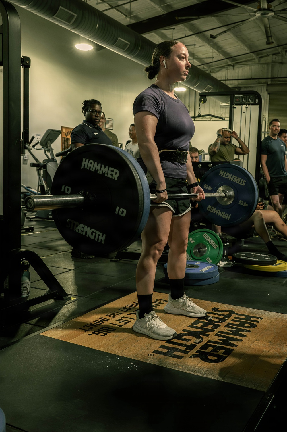 A woman is performing a deadlift in a gym, holding a loaded barbell at hip height in a completed lift position. She is standing on a Hammer Strength branded lifting platform, wearing a dark t-shirt, shorts, black knee-high socks, white sneakers, a lifting belt, and wrist wraps with wireless earbuds. Several onlookers and other gym-goers are visible in the background.