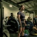 A woman is performing a deadlift in a gym, holding a loaded barbell at hip height in a completed lift position. She is standing on a Hammer Strength branded lifting platform, wearing a dark t-shirt, shorts, black knee-high socks, white sneakers, a lifting belt, and wrist wraps with wireless earbuds. Several onlookers and other gym-goers are visible in the background.