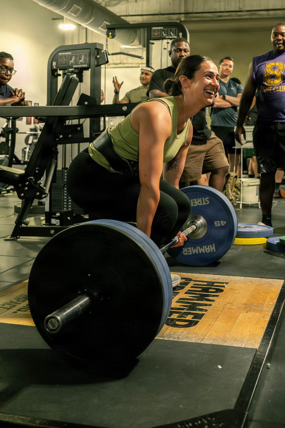 A woman in a green tank top and black leggings is performing a deadlift on a marked lifting platform in a gym, smiling during the lift. She is surrounded by a group of onlookers who appear to be cheering and watching her performance. The setting appears to be a structured gym environment with Hammer Strength branding visible on the platform and weight plates.