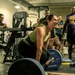 A woman in a green tank top and black leggings is performing a deadlift on a marked lifting platform in a gym, smiling during the lift. She is surrounded by a group of onlookers who appear to be cheering and watching her performance. The setting appears to be a structured gym environment with Hammer Strength branding visible on the platform and weight plates.