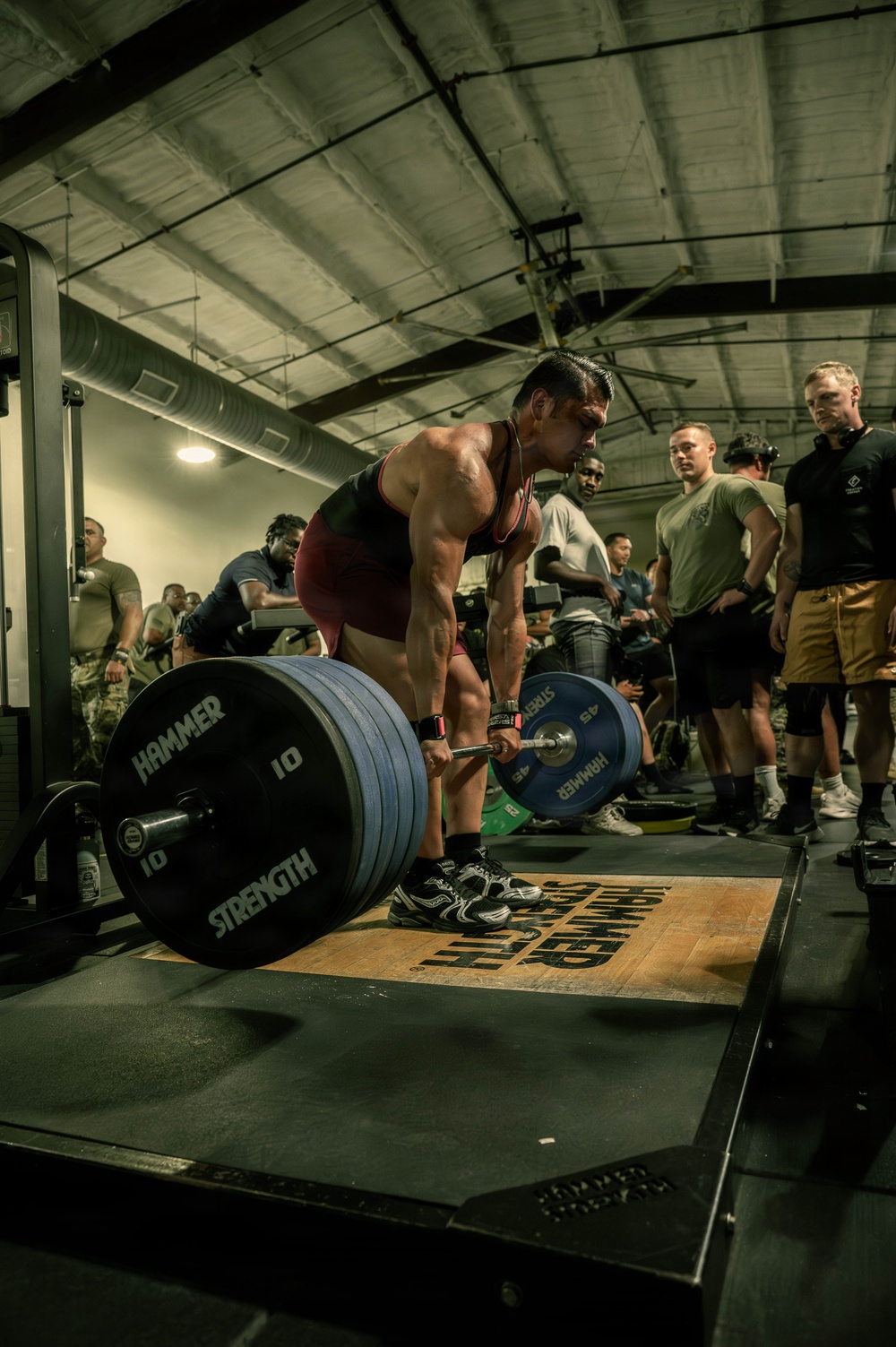 A muscular athlete in a red singlet performs a deadlift during what appears to be a competitive or organized weightlifting event in an indoor gym facility. The barbell is loaded with Hammer Strength plates and rests on a Hammer Strength branded lifting platform. Multiple spectators, several appearing to be in military attire, observe the lift from the sides.