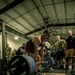 A muscular athlete in a red singlet performs a deadlift during what appears to be a competitive or organized weightlifting event in an indoor gym facility. The barbell is loaded with Hammer Strength plates and rests on a Hammer Strength branded lifting platform. Multiple spectators, several appearing to be in military attire, observe the lift from the sides.