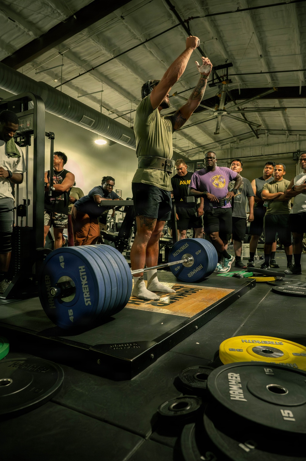 A weightlifter is performing what appears to be a snatch or overhead lift in a gym setting, with arms fully extended overhead. The lifter stands on a Hammer Strength platform surrounded by heavily loaded barbells with 45 lb plates. A crowd of approximately 10-15 spectators watches from behind, suggesting a competitive or demonstration event.
