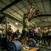 A weightlifter is performing what appears to be a snatch or overhead lift in a gym setting, with arms fully extended overhead. The lifter stands on a Hammer Strength platform surrounded by heavily loaded barbells with 45 lb plates. A crowd of approximately 10-15 spectators watches from behind, suggesting a competitive or demonstration event.