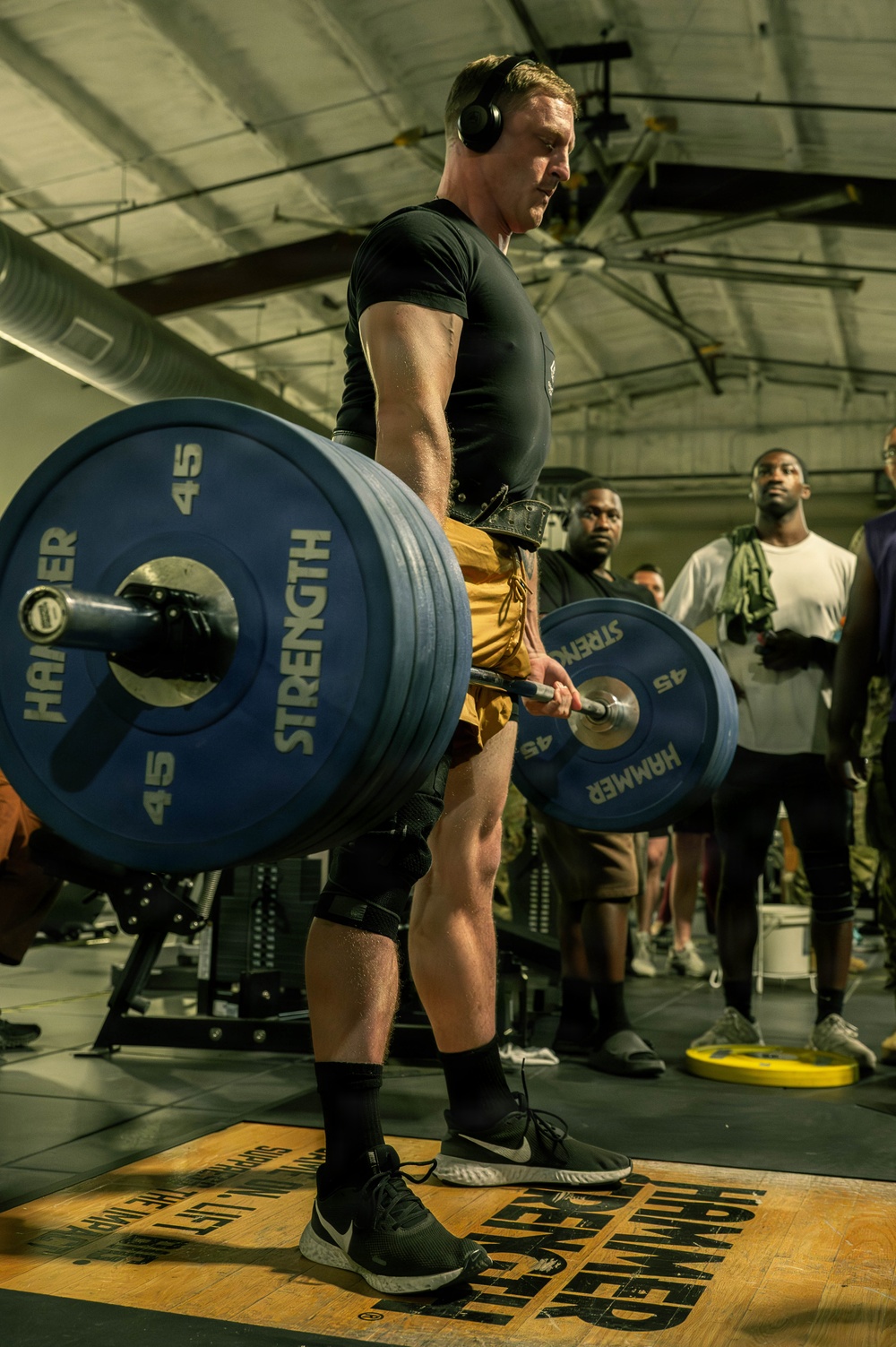 A male athlete is performing a deadlift in what appears to be a gym competition or training event, lifting a heavily loaded barbell with multiple 45-lb Hammer Strength plates. He is wearing a black compression shirt, yellow shorts, knee sleeves, a lifting belt, and over-ear headphones. A crowd of spectators is visible in the background observing the lift.