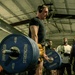 A male athlete is performing a deadlift in what appears to be a gym competition or training event, lifting a heavily loaded barbell with multiple 45-lb Hammer Strength plates. He is wearing a black compression shirt, yellow shorts, knee sleeves, a lifting belt, and over-ear headphones. A crowd of spectators is visible in the background observing the lift.