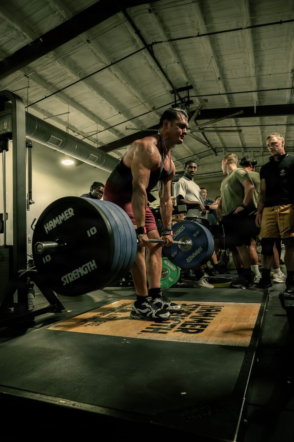 A muscular athlete is performing a deadlift in what appears to be a military or competitive gym setting, wearing a red tank top and shorts. The lifter is mid-lift with a heavily loaded barbell featuring Hammer Strength bumper plates. Several spectators, many appearing to be in military attire, observe in the background.