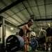 A muscular athlete is performing a deadlift in what appears to be a military or competitive gym setting, wearing a red tank top and shorts. The lifter is mid-lift with a heavily loaded barbell featuring Hammer Strength bumper plates. Several spectators, many appearing to be in military attire, observe in the background.